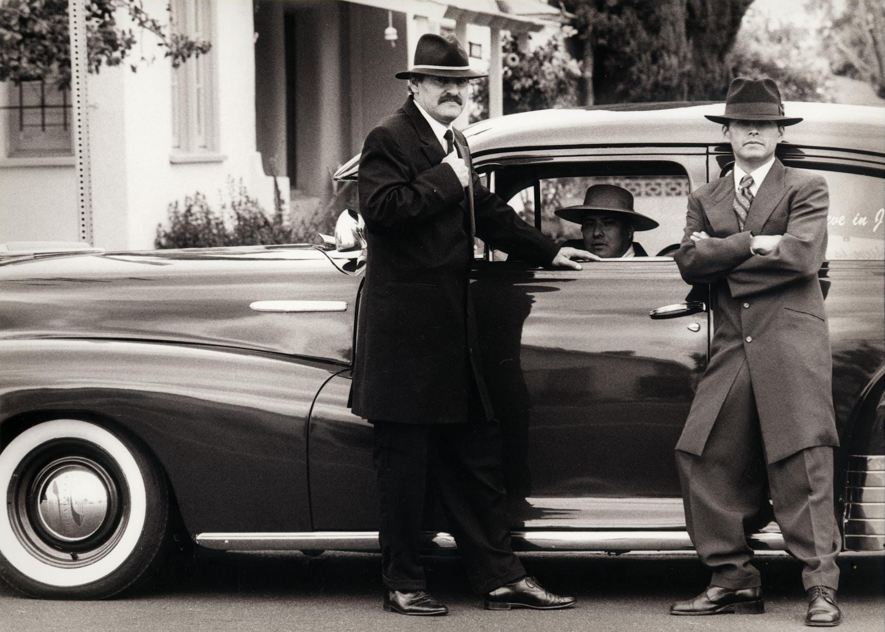 Três homens com ternos vintage e chapéus fedora estão ao lado de um clássico carro dos anos 1930 em frente a um edifício estilo Chicago, foto em preto e branco.