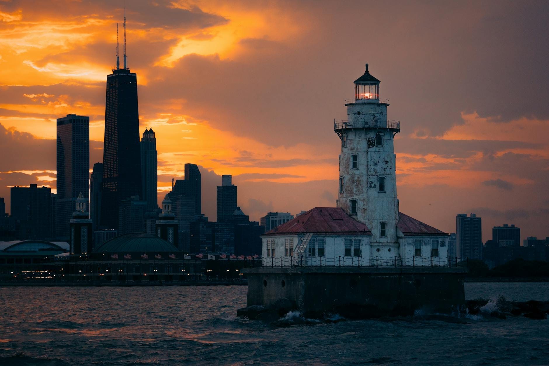 Il Faro del Porto di Chicago illuminato al tramonto con lo skyline della città e un cielo arancione intenso sullo sfondo, visto dall'acqua.
