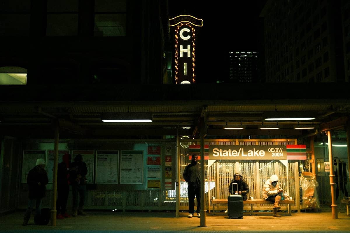 Nächtliche Szene am erhöhten Bahnhof State/Lake in Chicago mit wartenden Menschen, beleuchteter Beschilderung und einem klassischen Chicago-Theaterschild im Hintergrund.