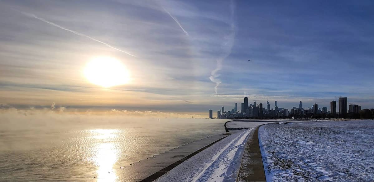 Verschneiter Uferweg am See in Chicago mit der Skyline der Stadt und aufsteigendem Nebel über dem Lake Michigan im Winterlicht.