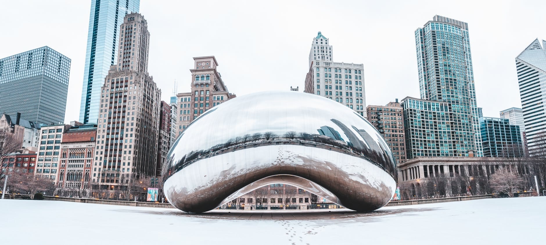 Vista ampla da escultura Cloud Gate no Millennium Park de Chicago, cercada por neve no chão e arranha-céus ao fundo em um dia frio e claro de inverno.