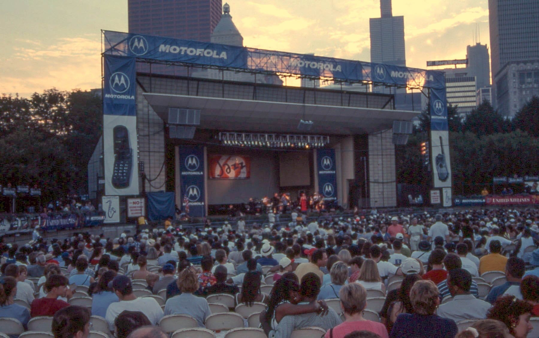 Grande folla che si gode un'esibizione jazz dal vivo sul palco all'aperto del Millennium Park a Chicago con i grattacieli al tramonto.