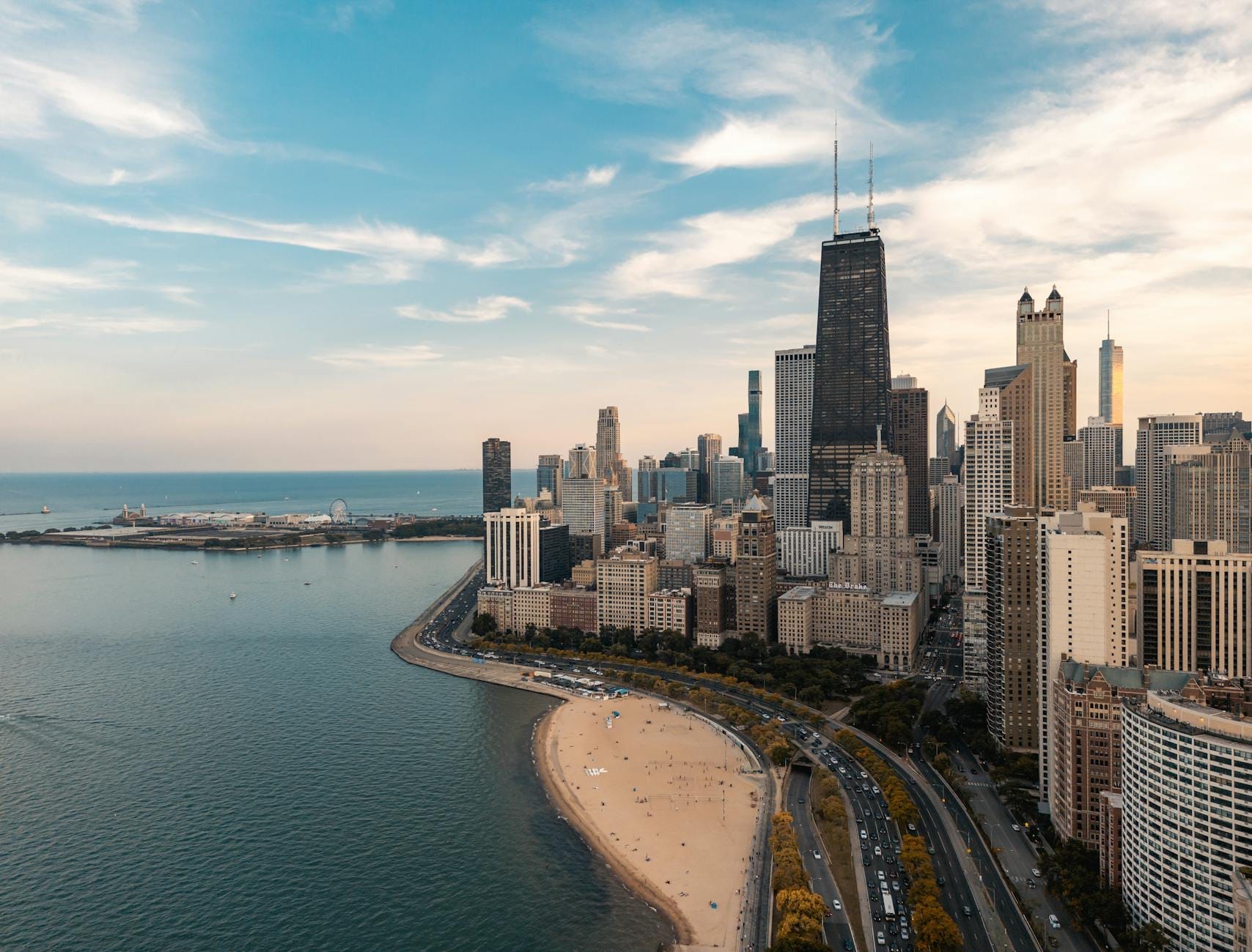 Vista aérea da orla de Chicago com praia de areia, área verde e o skyline do centro sob um céu azul ao pôr do sol.