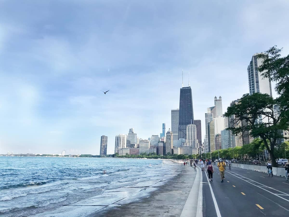 Sendero pavimentado junto al lago Michigan con personas caminando y en bicicleta, horizonte de Chicago visible al fondo bajo un cielo azul.