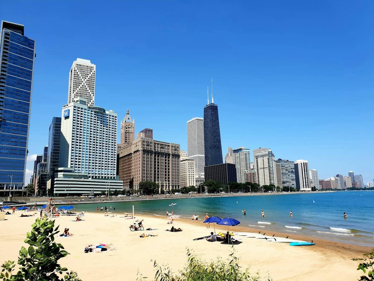 Playa de arena en el lago de Chicago con personas tomando el sol y nadando, rascacielos y cielo azul al fondo.