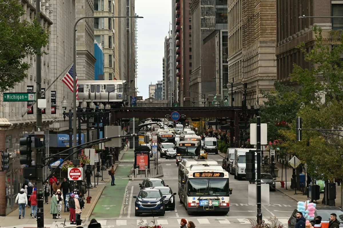 Affollata strada del centro di Chicago con autobus CTA, auto, pedoni e un treno 'L' che passa in sopraelevata vicino a Michigan Avenue.