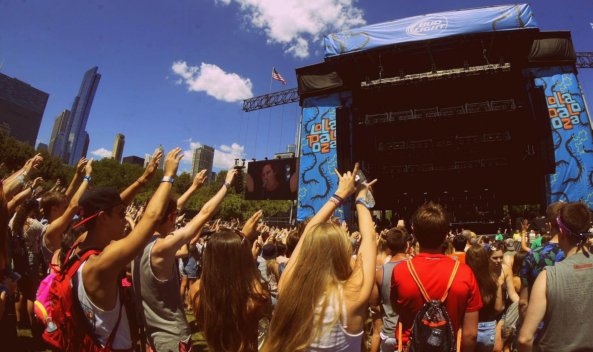 Grande multidão reunida em frente ao palco do Lollapalooza em Grant Park, Chicago, com os arranha-céus da cidade e um céu azul brilhante ao fundo.