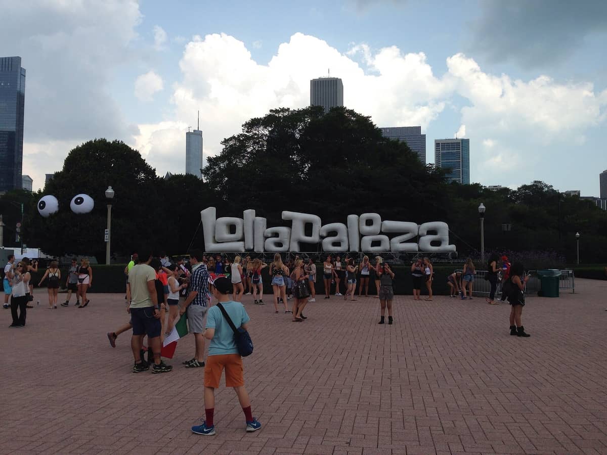 I visitatori si radunano davanti a un grande cartello bianco di Lollapalooza nel Grant Park, Chicago, con grattacieli sullo sfondo e nuvole estive in alto.