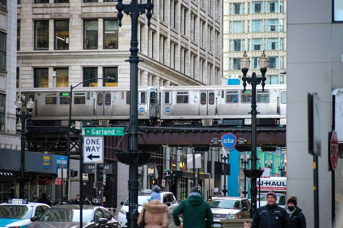 Chicago CTA 'L' train passing overhead in downtown, with visible CTA sign, city street, and people below on a clear day.