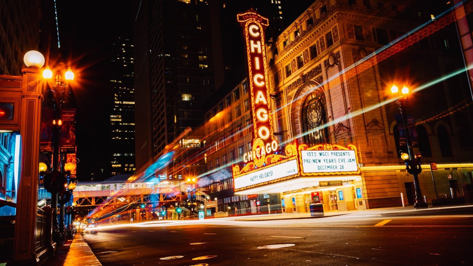 Cena noturna vibrante do icônico Chicago Theatre, com letreiros de néon acesos, rastros de luz de carros e arranha-céus ao fundo.