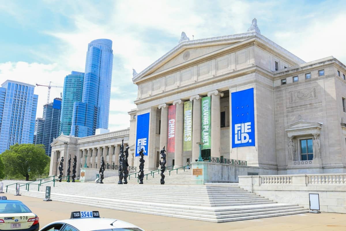 Das Field Museum in Chicago an einem sonnigen Tag, mit Bannern, breiten Treppen und Wolkenkratzern der Stadt im Hintergrund.