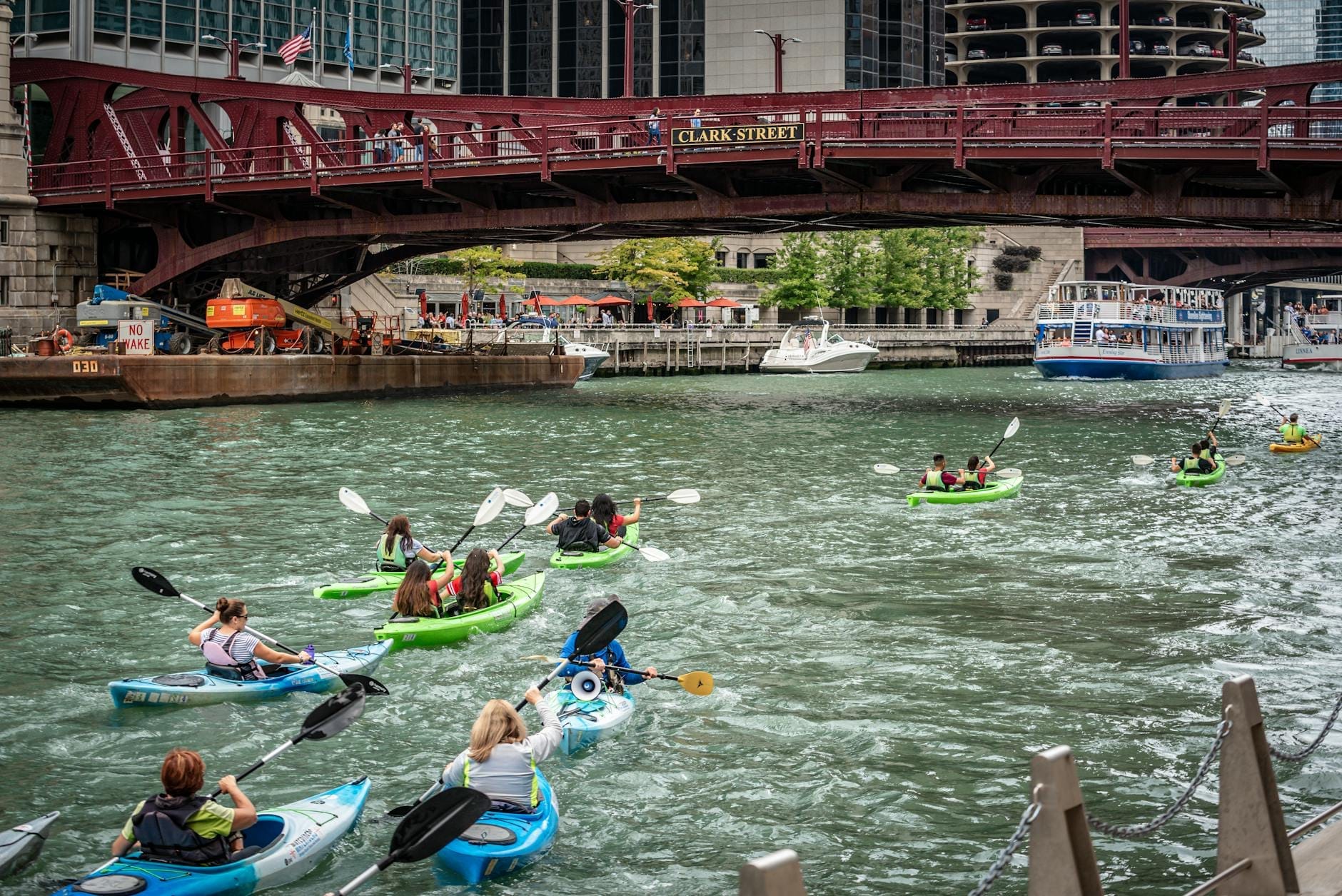 Un gruppo di kayaker pagaia sul Chicago River sotto un ponte rosso, con grattacieli e una barca turistica sullo sfondo.