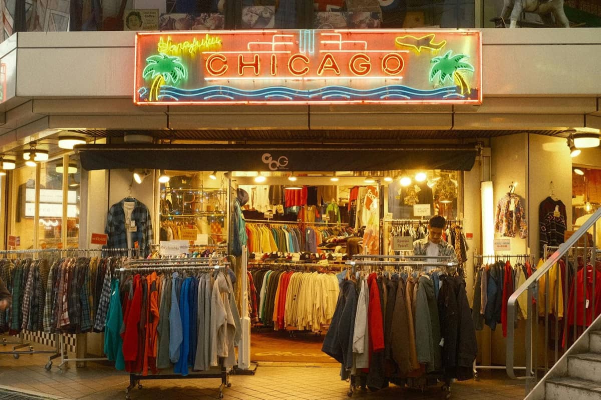 Ladenfront mit Kleiderständern draußen, Neonschild mit der Aufschrift 'Chicago' über dem Eingang und eine Person, die drinnen stöbert.