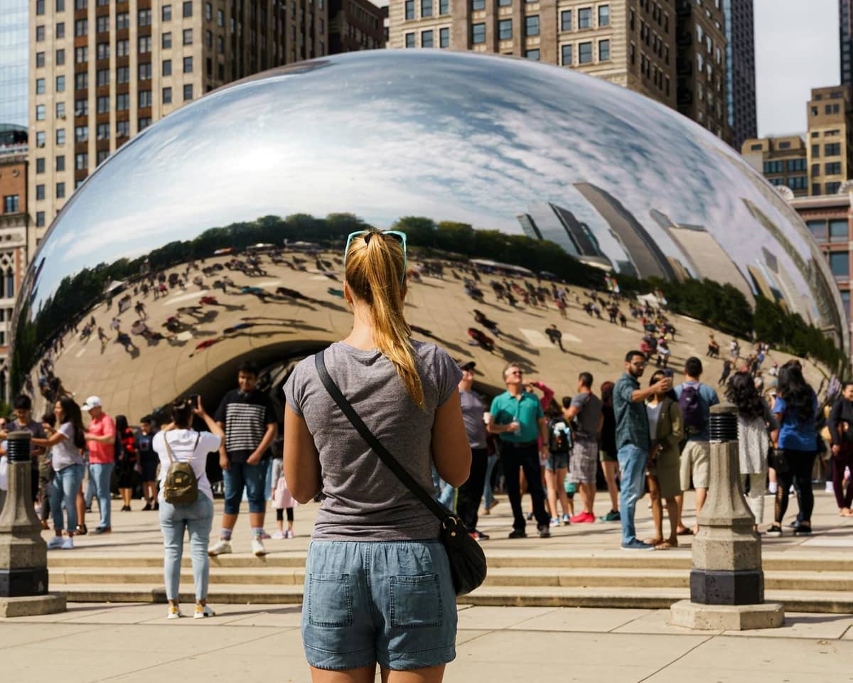 Ein Alleinreisender steht vor Chicagos Cloud Gate (The Bean) inmitten einer lebhaften Menge, umgeben von Wolkenkratzern der Innenstadt an einem sonnigen Tag.