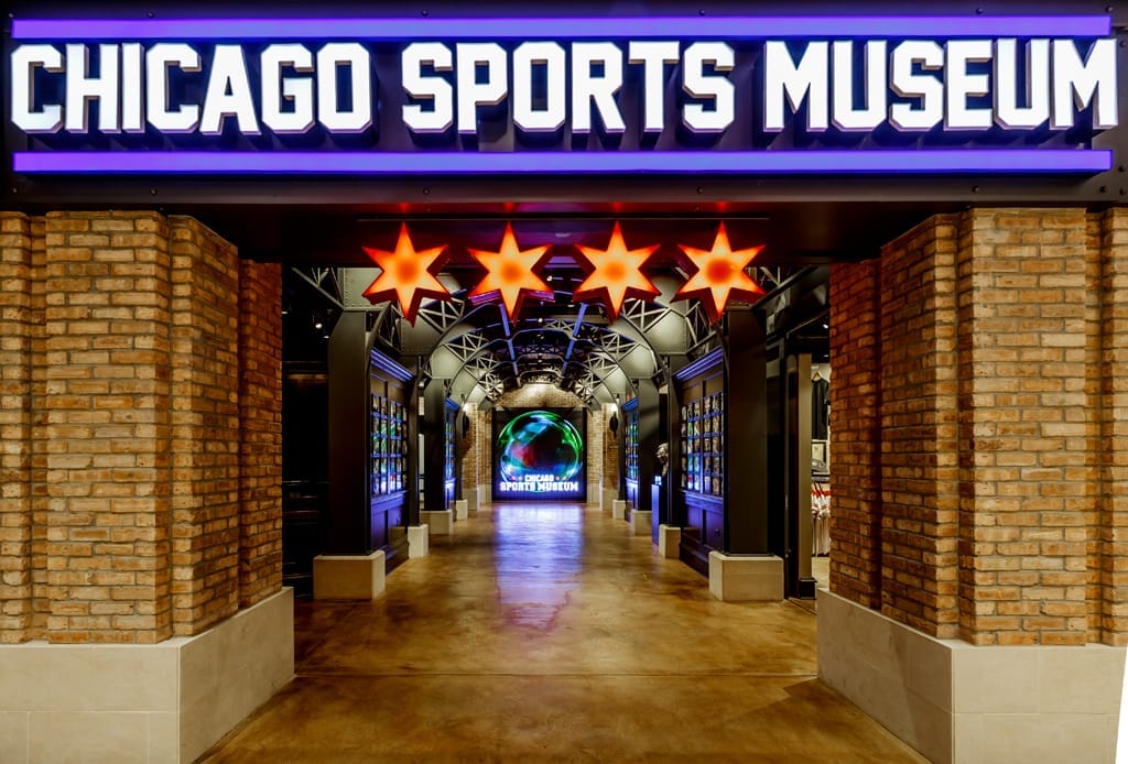 Ingresso al Chicago Sports Museum con insegna illuminata, stelle luminose di Chicago, pareti in mattoni a vista e display sportivi lungo il corridoio.