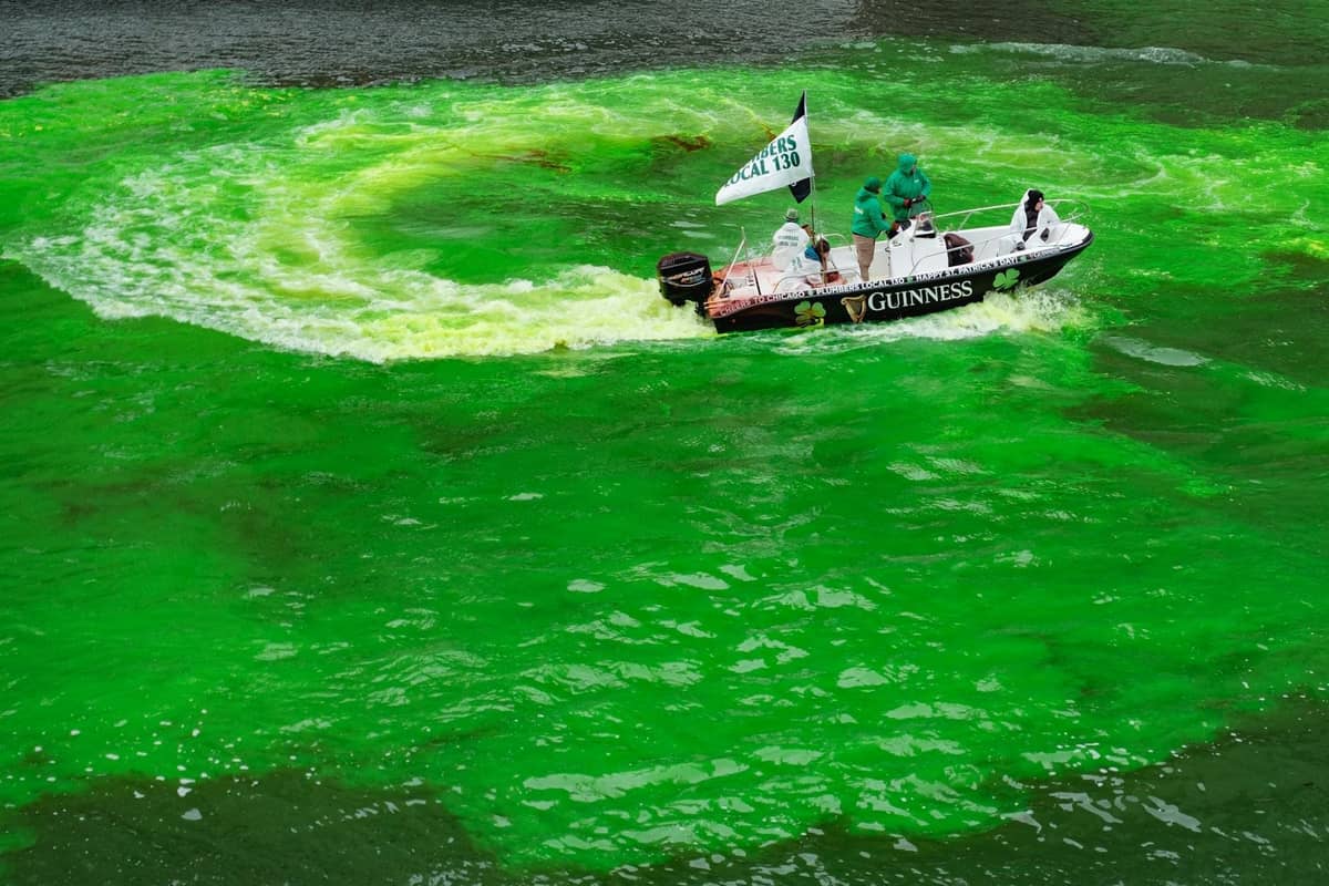 Boat with people in festive attire actively dyeing the Chicago River bright green, swirling water during St. Patrick's Day celebration.