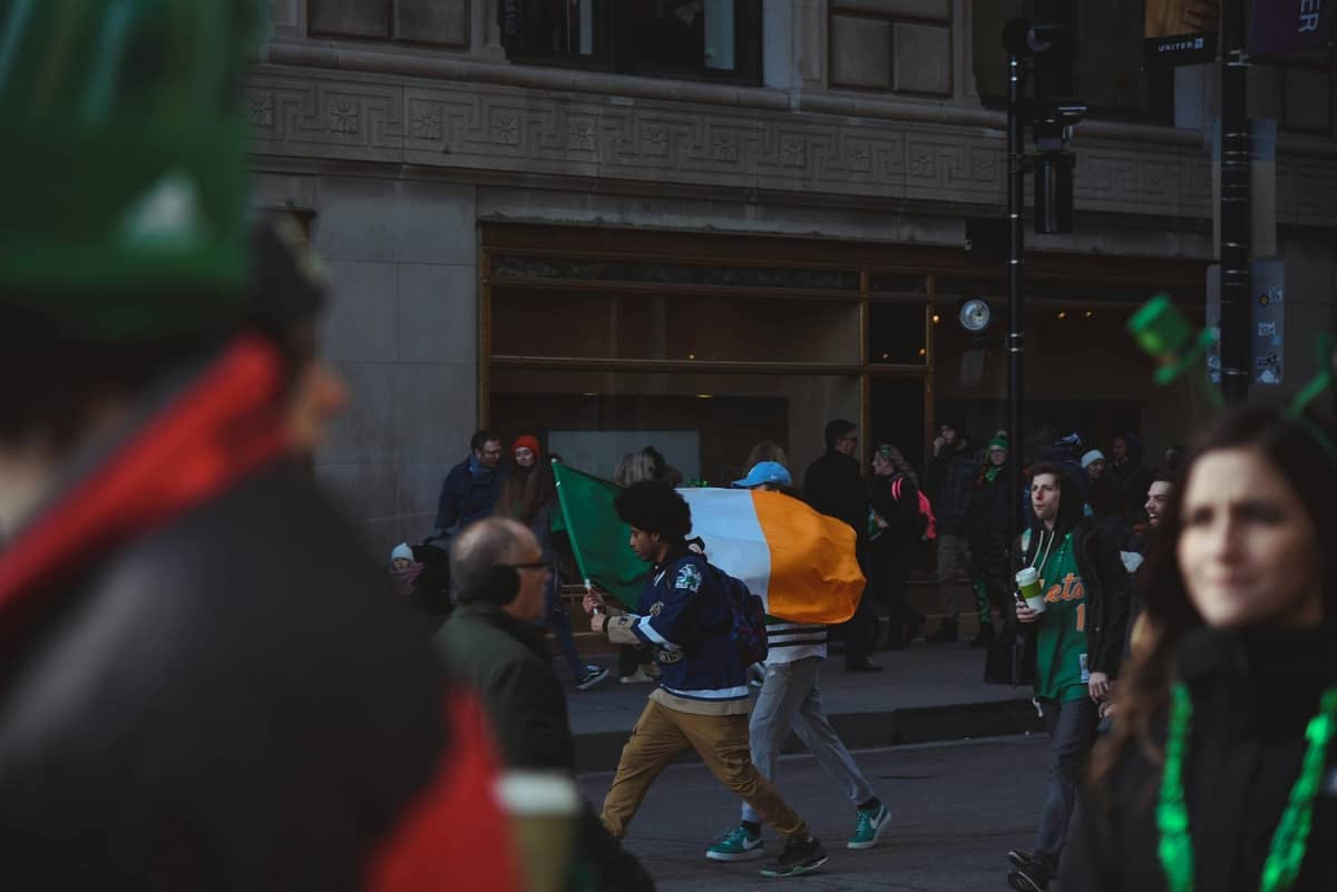 Crowd celebrating St. Patrick's Day outdoors, with a man carrying an Irish flag along a city street, buildings in background.