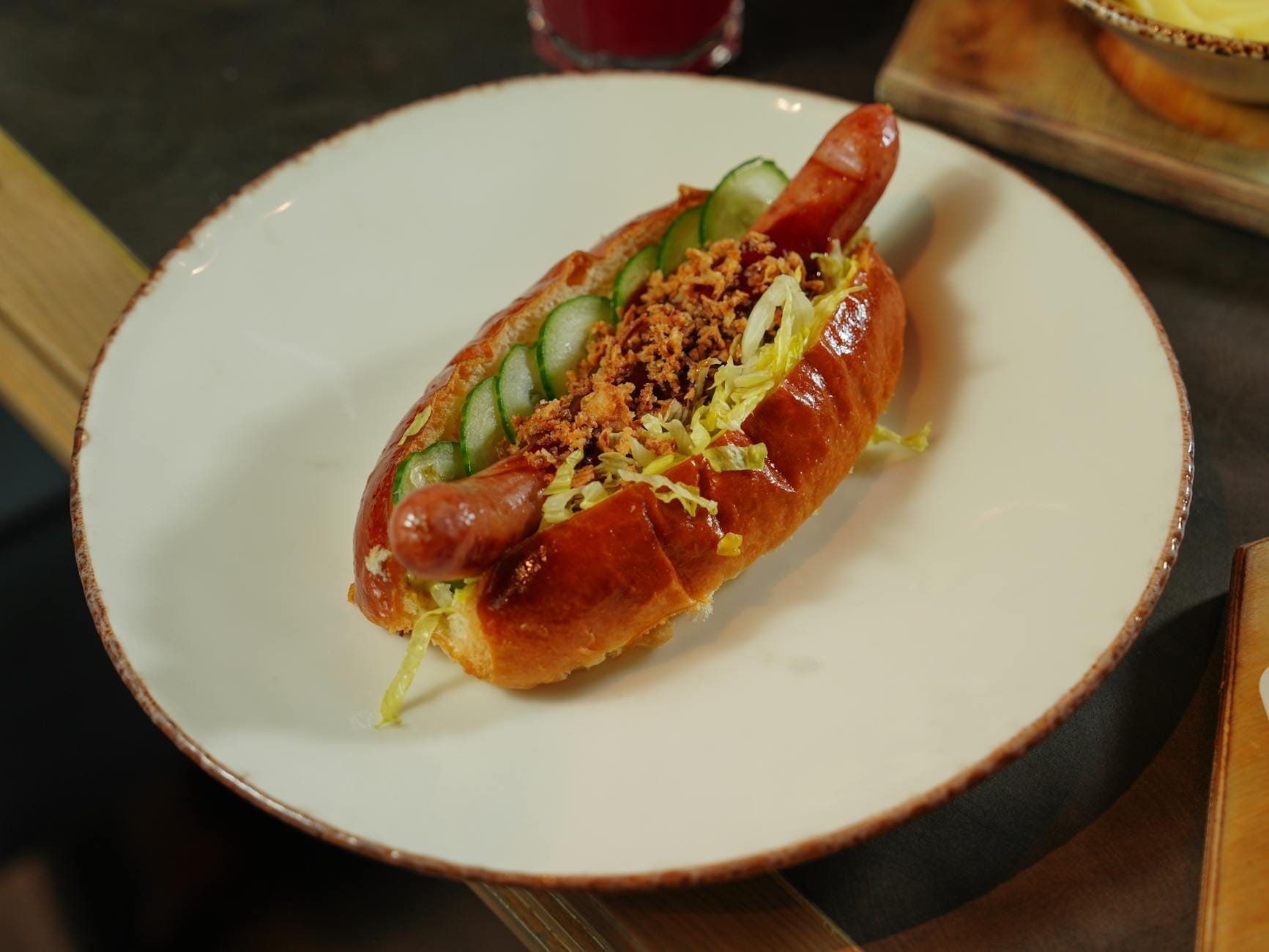 Um cachorro-quente estilo Chicago coberto com fatias de pepino, alface desfiada e pedaços crocantes em um prato branco com iluminação interna suave.
