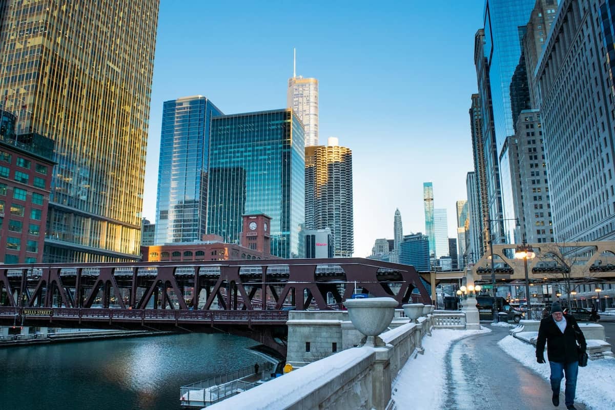 Vista del río Chicago, un puente elevado y el Riverwalk con rascacielos que reflejan el sol de la mañana. Una persona camina por el sendero nevado junto al río en invierno.