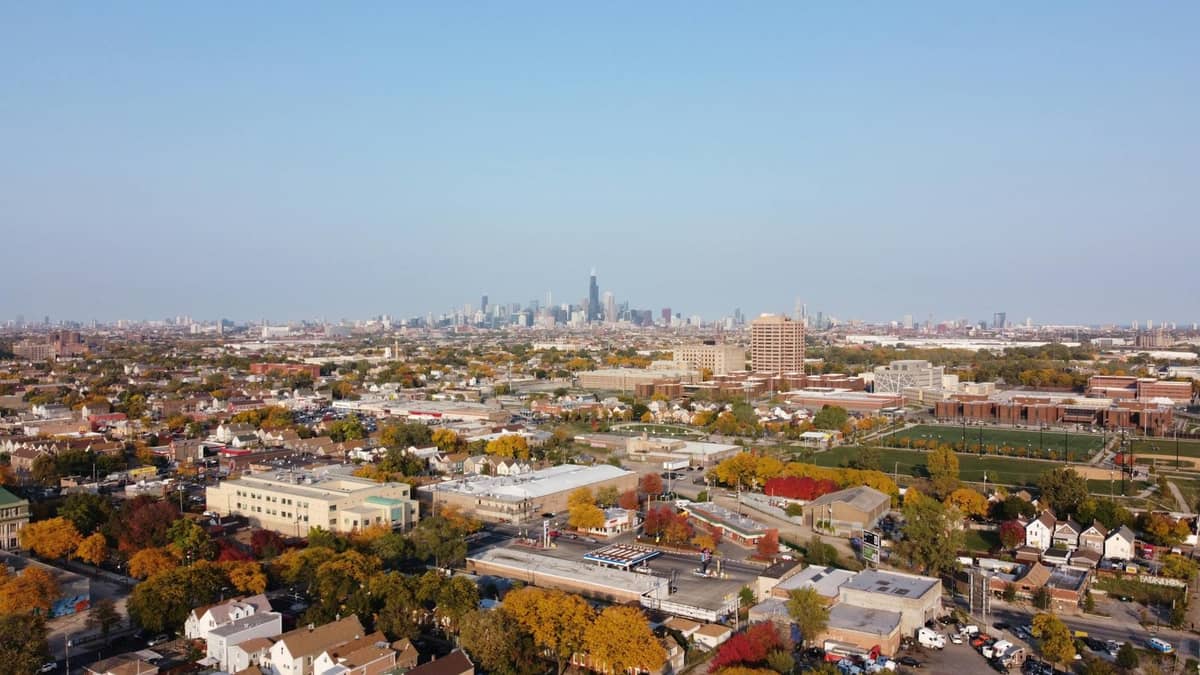 Vista aérea amplia de un vecindario de Chicago con casas y árboles en primer plano, y el horizonte del centro de Chicago a lo lejos bajo un cielo azul despejado.