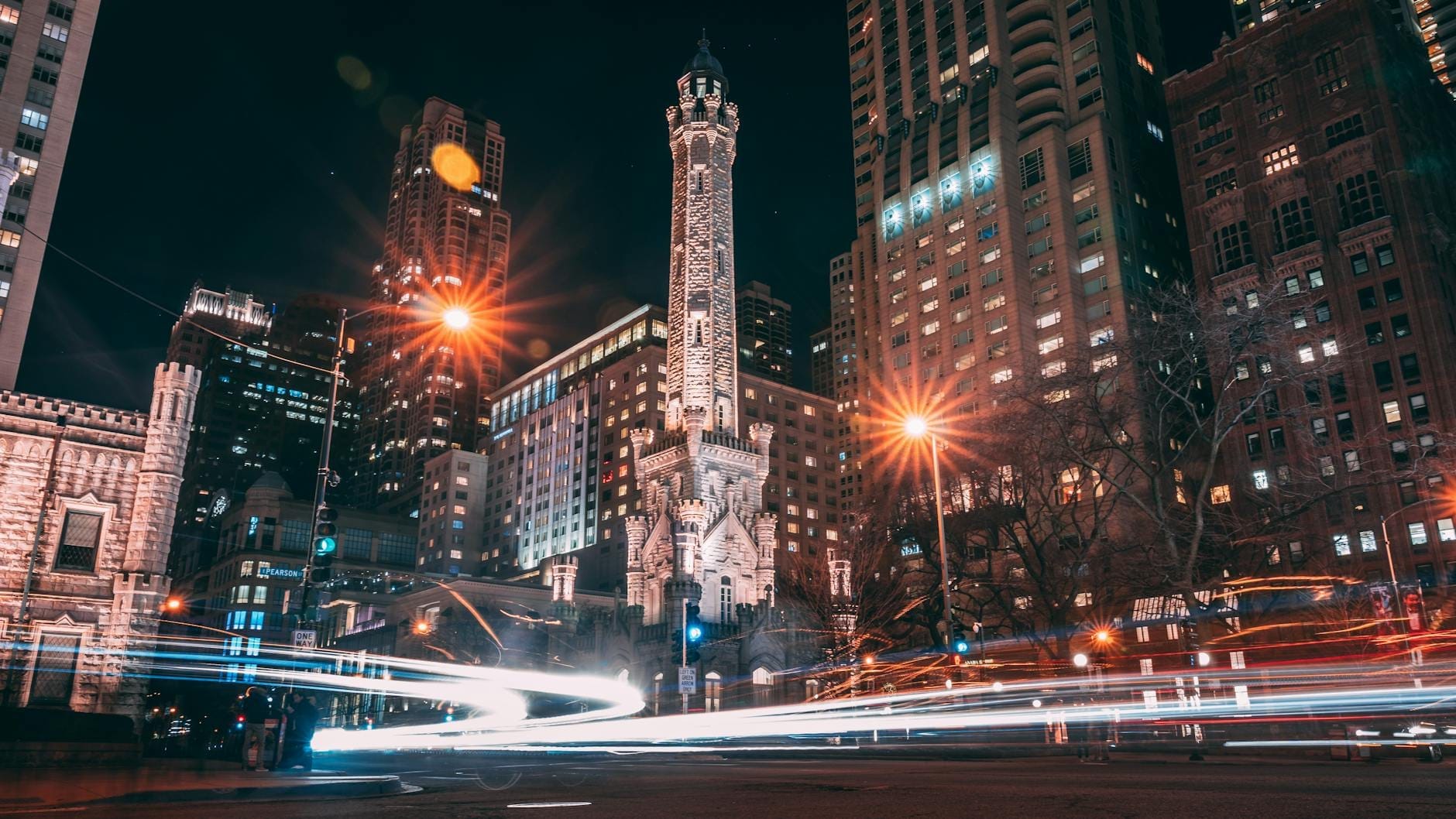 Vista notturna della Chicago Water Tower illuminata, circondata dai grattacieli moderni su North Michigan Avenue, con le scie luminose delle auto che aggiungono dinamismo alla scena.