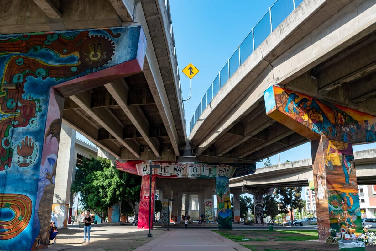 Unter der San Diego–Coronado Bridge im Chicano Park bedecken lebendige Wandgemälde mit farbenfrohen Chicano- und mexikanisch-amerikanischen Motiven die Betonpfeiler, während Menschen unter der Brücke entlanggehen.
