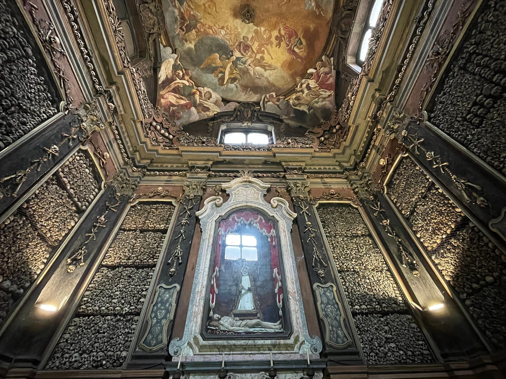 Interno della cappella ossario della Chiesa di San Bernardino alle Ossa, con pareti ricoperte di teschi e ossa umane e un soffitto barocco dipinto.
