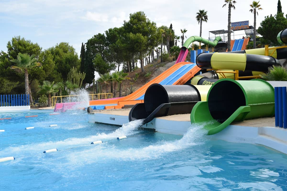 Colorful water slides emerge from a lush hillside into a large blue pool.