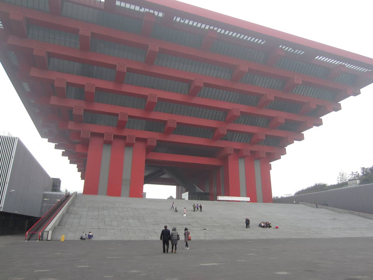 Weitwinkelperspektive auf das mächtige rote Gebäude des China Art Museum in Shanghai mit seiner beeindruckenden Architektur und dem Haupteingang, dem Besucher entgegengehen.