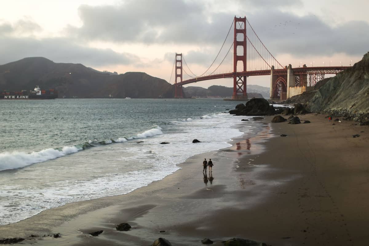 Vista di China Beach a San Francisco con il Golden Gate Bridge sullo sfondo, onde gentili, scogliere rocciose e alcune persone che camminano lungo la riva.