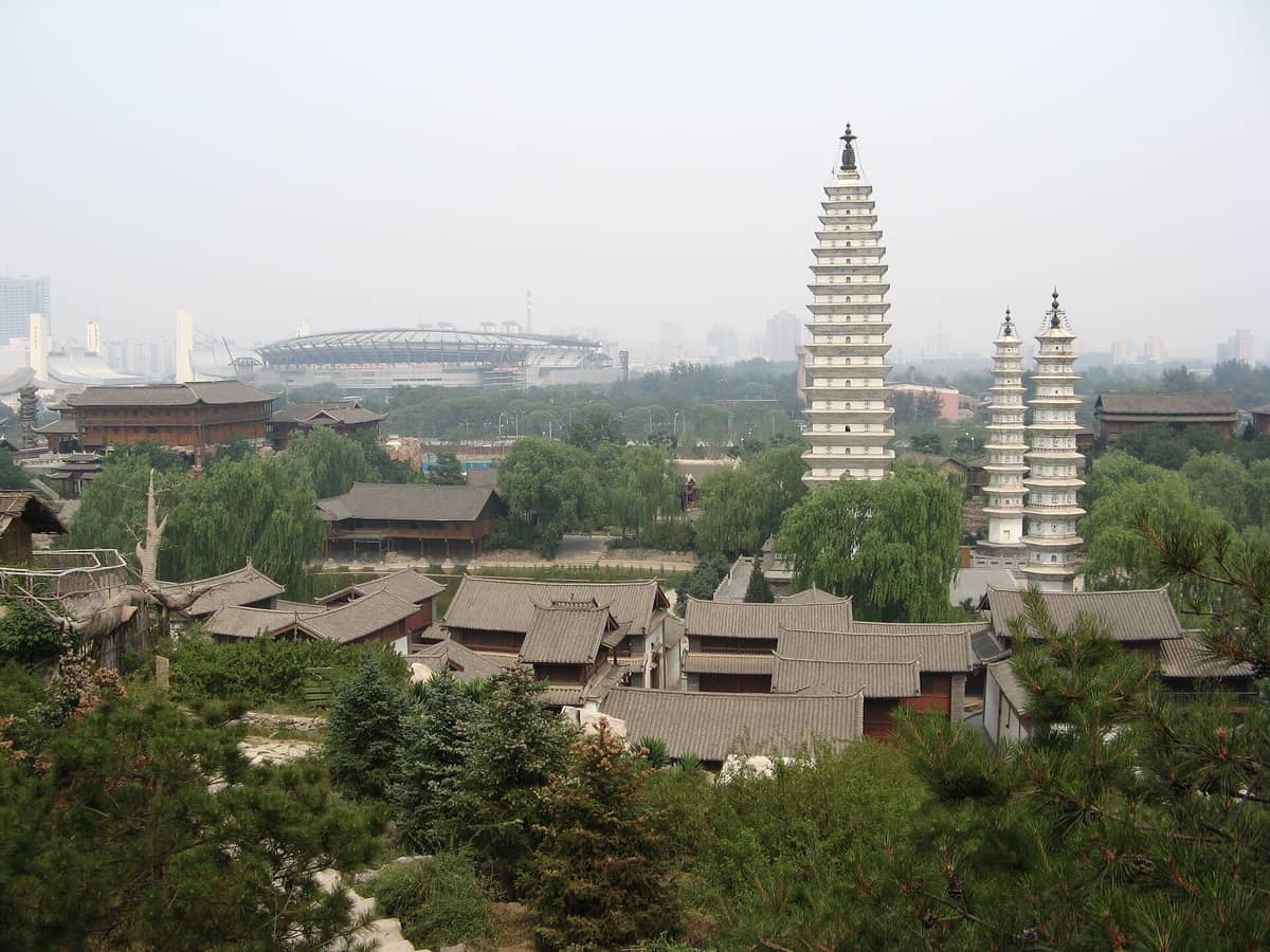 Blick auf den Chinesischen Volkskulturpark in Beijing mit traditionellen grau gedeckten Gebäuden, weißen Pagoden und üppigen grünen Bäumen an einem diesigen Tag.