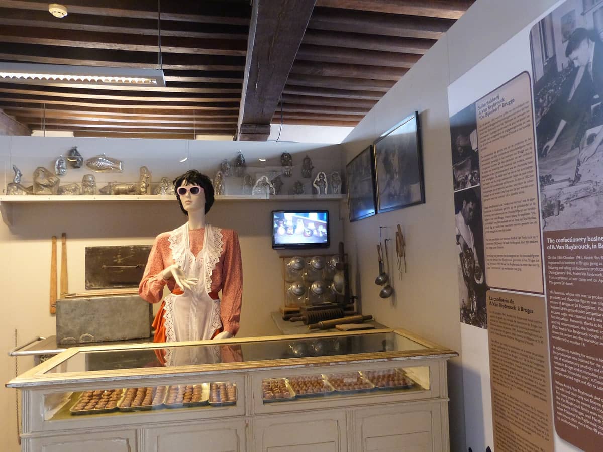 Display of vintage chocolate molds, historic tools, and a mannequin in period dress at Choco-Story Bruges, with trays of pralines in a glass case.