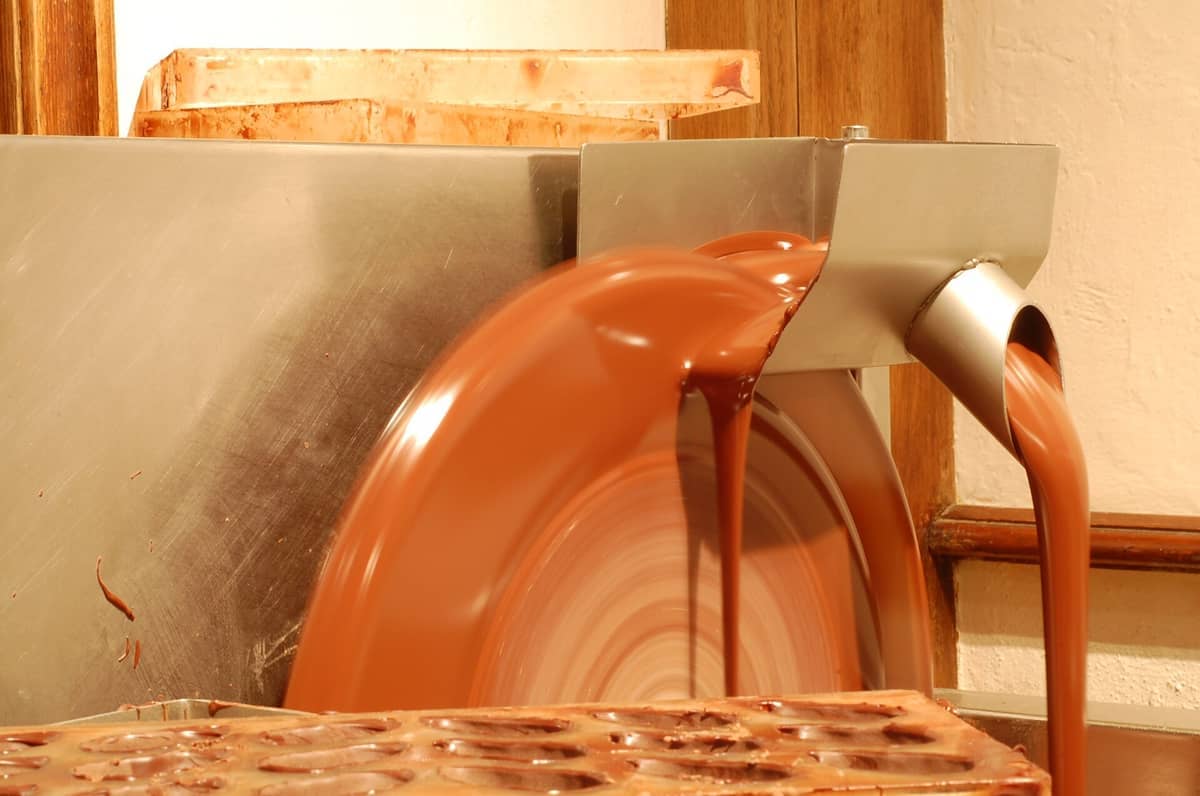 Close-up de chocolate derretido fluindo sendo processado por uma máquina no Choco-Story Bruxelas, com iluminação quente e detalhes de madeira visíveis.