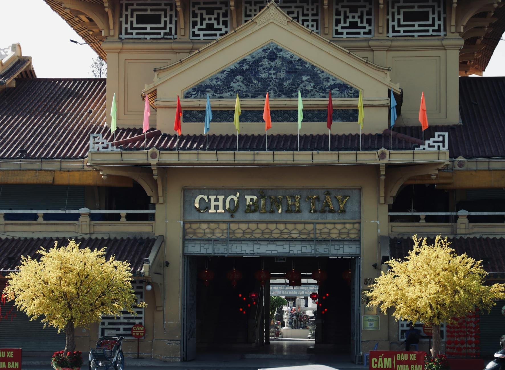 Vista frontal del histórico mercado Chợ Bình Tây en Chợ Lớn, adornado con banderas de colores y árboles con flores amarillas, una invitación a explorar el barrio chino.