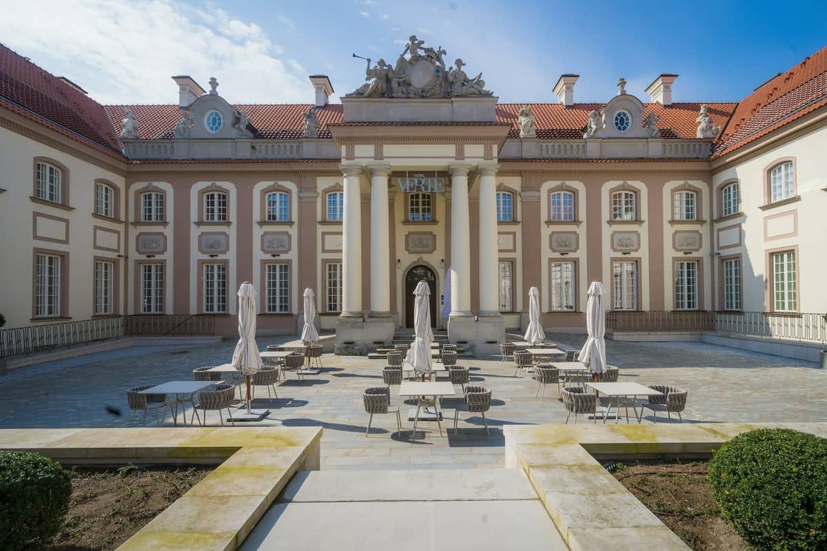 Elegancki dziedziniec barokowego pałacu ze stolikami kawiarni, parasolami i ozdobnymi rzeźbami nad wejściem, pod błękitnym niebem w Warszawie.