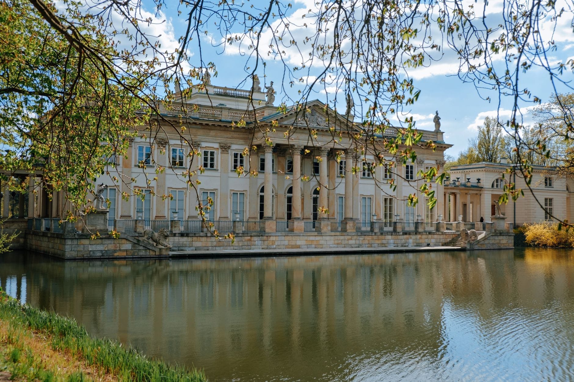 Palácio neoclássico à beira de um lago tranquilo no Parque Łazienki de Varsóvia, emoldurado por galhos de árvores e céu azul, convidando os amantes da música a explorar o legado de Chopin.
