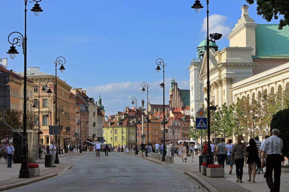 Szeroki widok na Krakowskie Przedmieście w Warszawie, otoczone historycznymi budynkami i tętniące życiem przechodniów w słoneczny dzień.