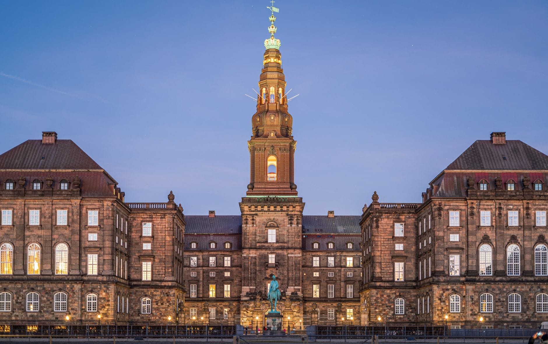 Il Palazzo di Christiansborg al tramonto con la torre centrale illuminata e una statua in primo piano, visti da un'ampia piazza aperta sotto un cielo sereno color blu serale.