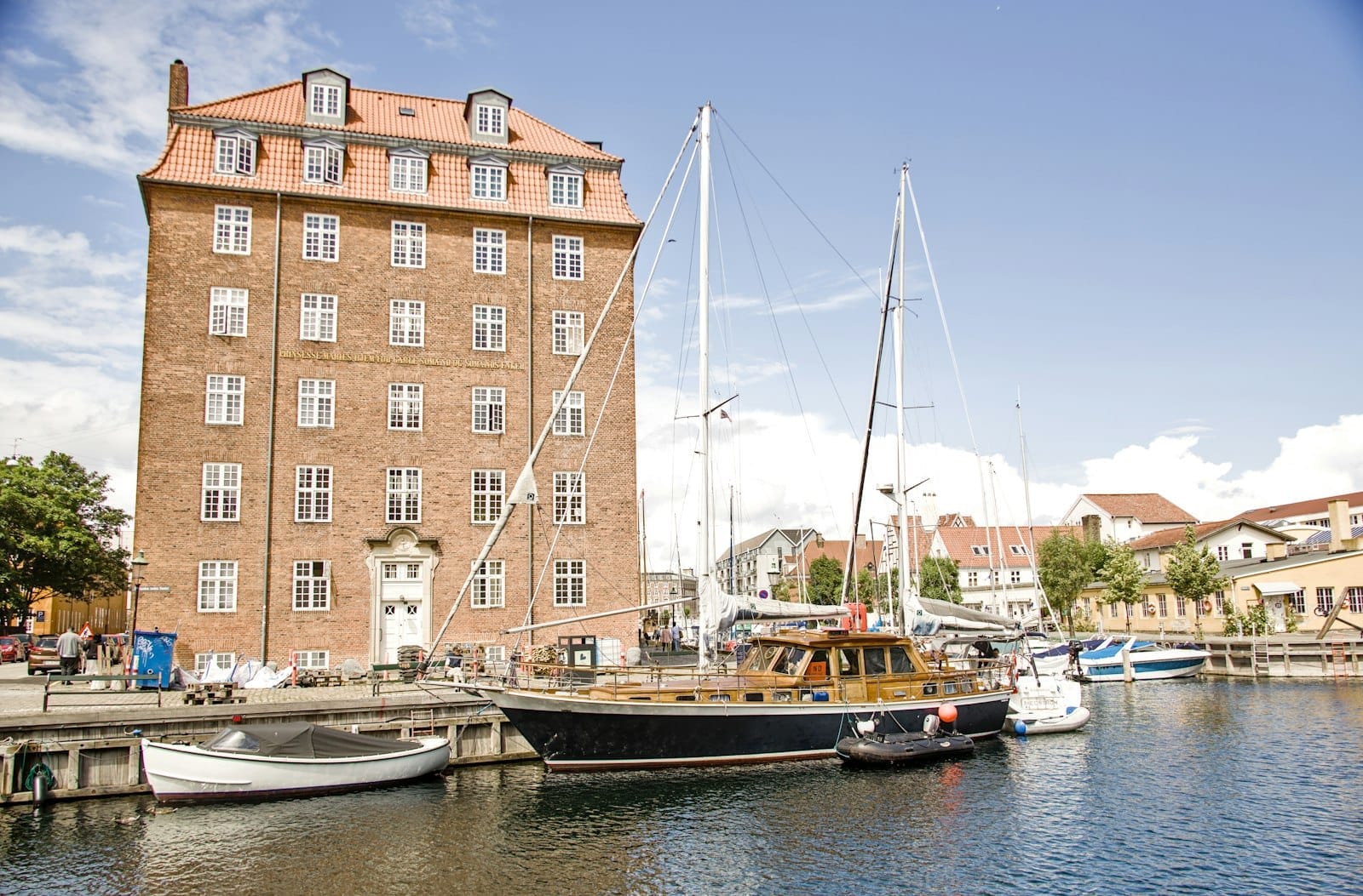อาคารอิฐประวัติศาสตร์ที่มีกระเบื้องหลังคาสีแดงเรียงรายตามคลอง Christianshavn พร้อมเรือใบและเรือขนาดเล็กจอดเทียบท่าในสภาพอากาศแดดจ้า