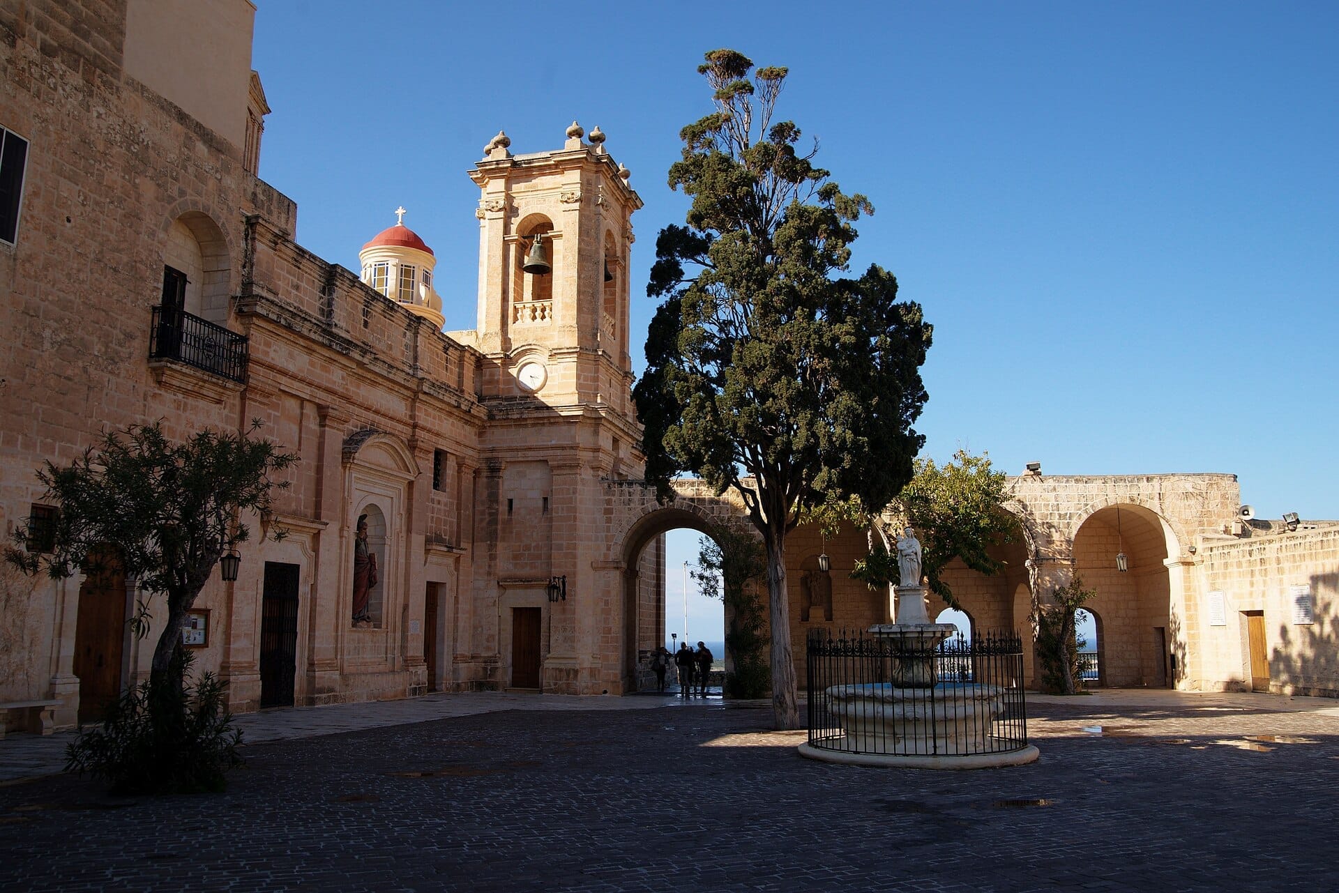 Cortile del Santuario di Nostra Signora di Mellieħa con muri in pietra calcarea, campanile, ingresso ad arco, alberi, una statua centrale e cielo azzurro limpido.