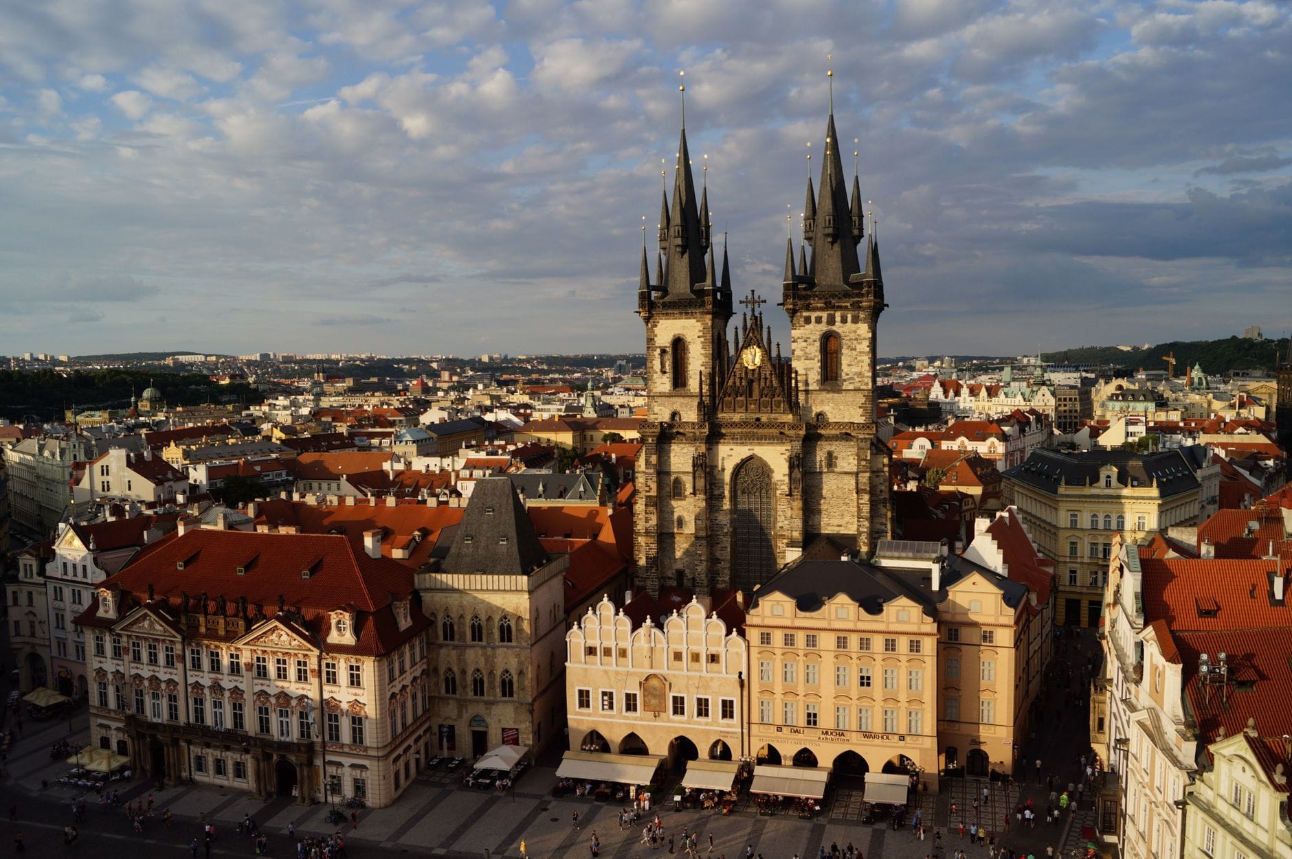 Vista grandangolare delle due guglie gotiche della Chiesa di Nostra Signora di Týn che si innalzano sulla Piazza della Città Vecchia di Praga, con i tetti rossi degli edifici sotto un cielo parzialmente nuvoloso.