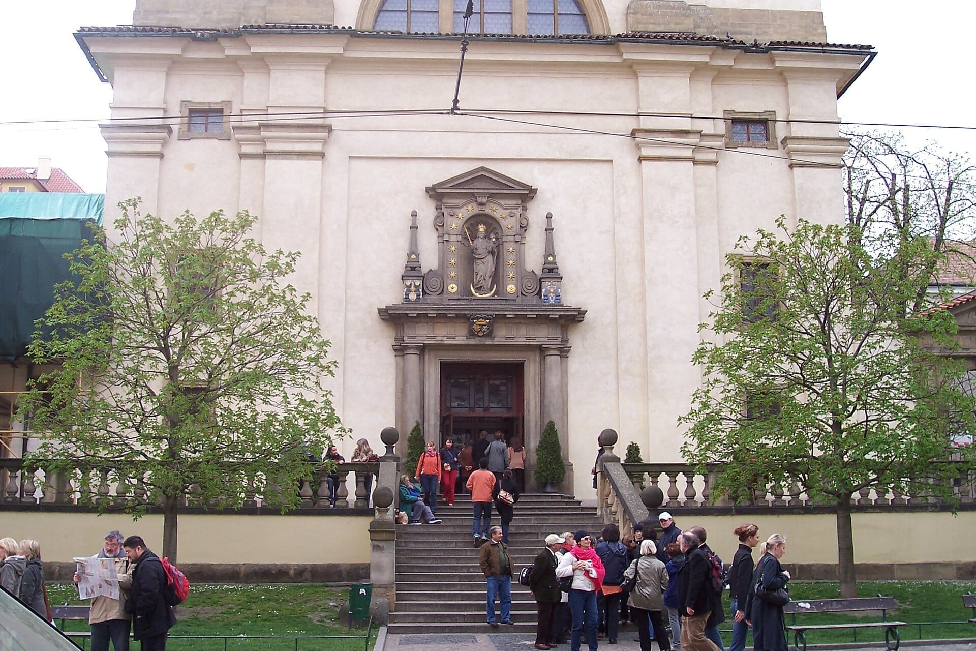 I visitatori si radunano all'ingresso della Chiesa di Nostra Signora della Vittoria a Praga, con la sua facciata barocca e una statua decorativa sopra il portale.