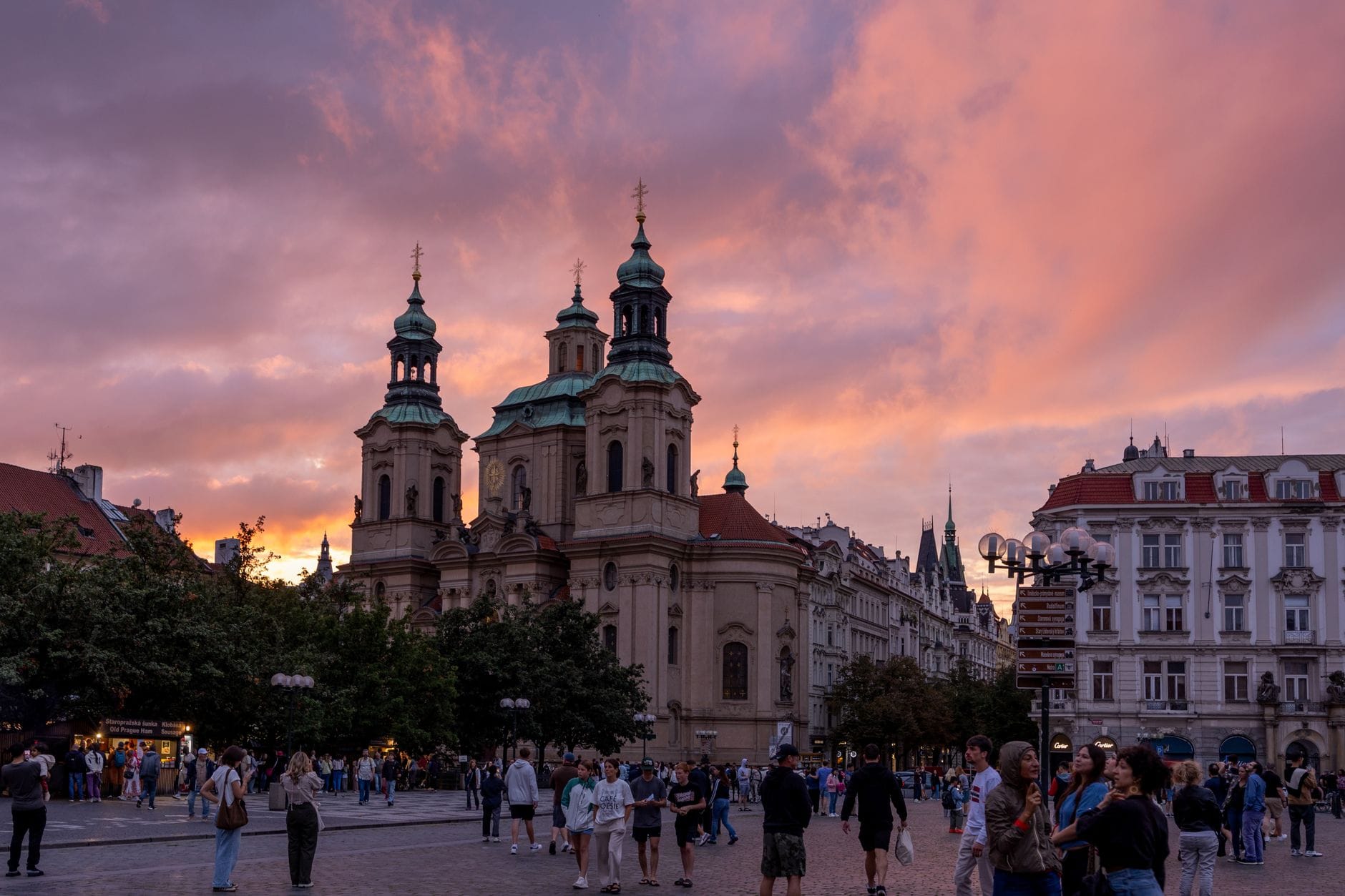 La Chiesa di San Nicola in Piazza della Città Vecchia di Praga al tramonto, con nuvole rosa acceso nel cielo e una folla animata davanti alla facciata barocca.