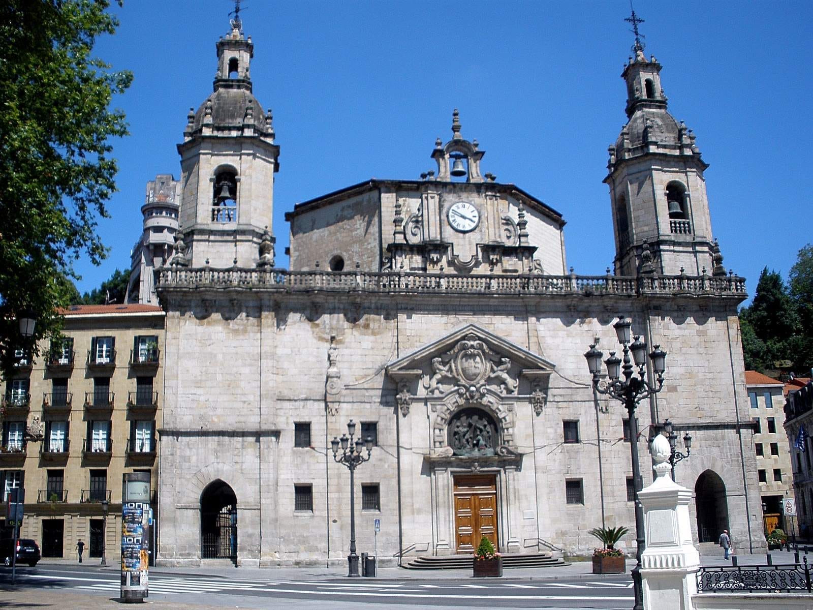 Facade of the Church of San Nicolás de Bari in Bilbao, featuring twin bell towers, ornate Baroque details, and a bright blue sky.