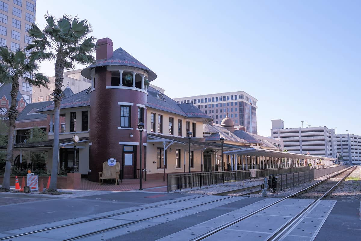 Wide view of Church Street Station’s historic brick and yellow railroad depot, with palm trees, train tracks, and downtown Orlando buildings in the background.