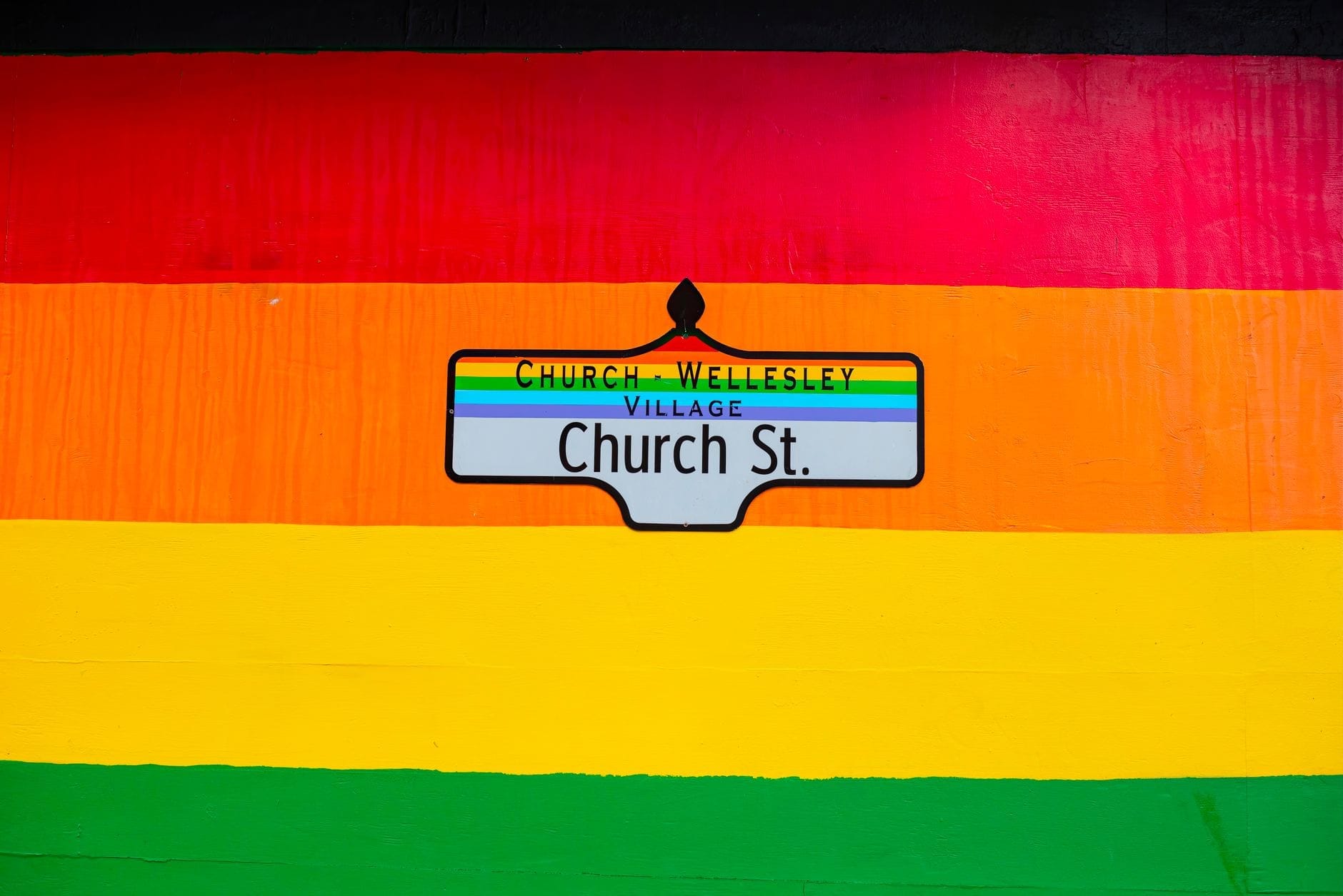ภาพกราฟิตีสีรุ้งสดใสพร้อมป้ายถนน Church-Wellesley Village อยู่ตรงกลาง เป็นสัญลักษณ์ของย่าน LGBTQ+ ในโตรอนโตที่เปี่ยมไปด้วยพลังและบรรยากาศอบอุ่น
