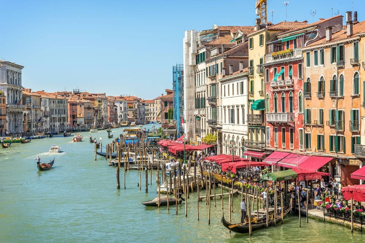 Tageslichtansicht des Canal Grande in Venedig mit bunten Gebäuden und roten Markisen am Wasser, belebt von Menschen und Gondeln, in der Nähe des Rialto-Markts.