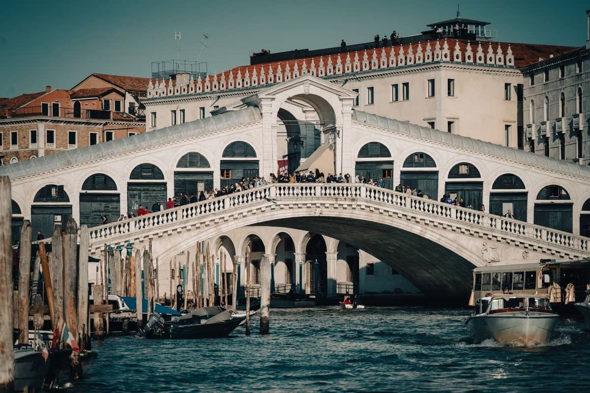 Weitwinkelansicht der belebten Rialtobrücke in Venedig mit Menschen, die darüber gehen, Booten, die darunter vorbeifahren, und historischen Gebäuden entlang des Kanals.