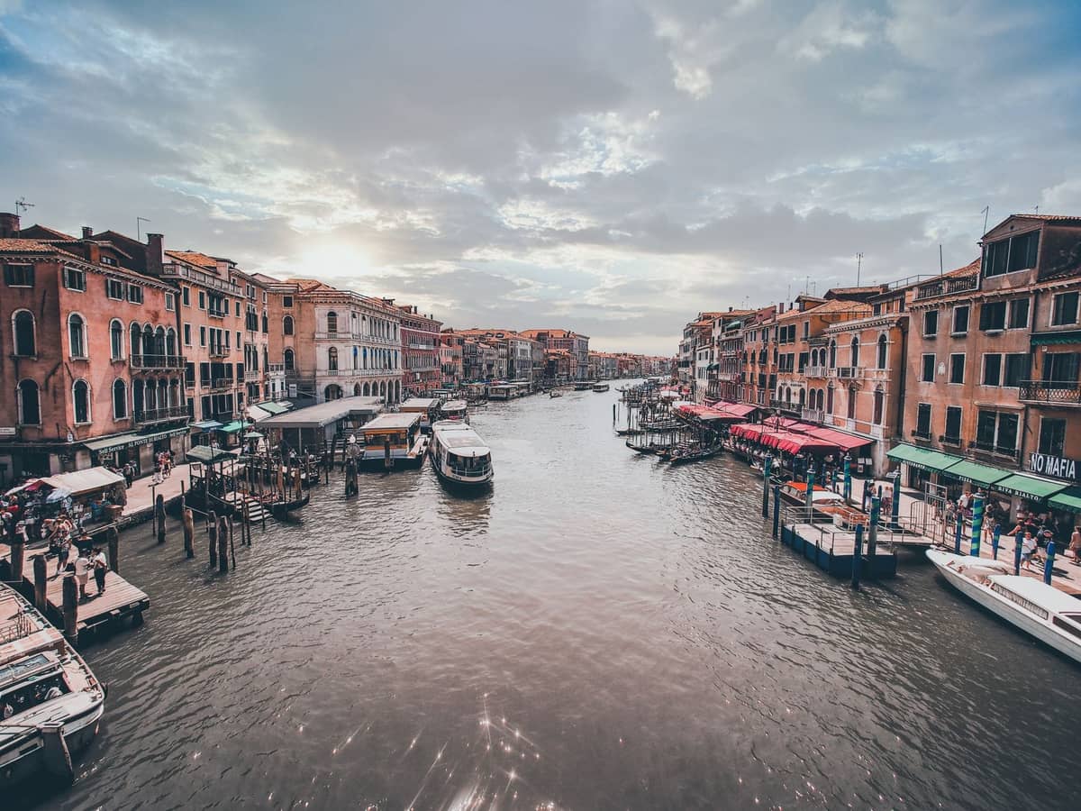 Weiter Blick auf den Canal Grande in Venedig mit bekannten Bacari-Terrassenbars, roten Markisen, Booten und Menschen an beiden Ufern in der Abenddämmerung.
