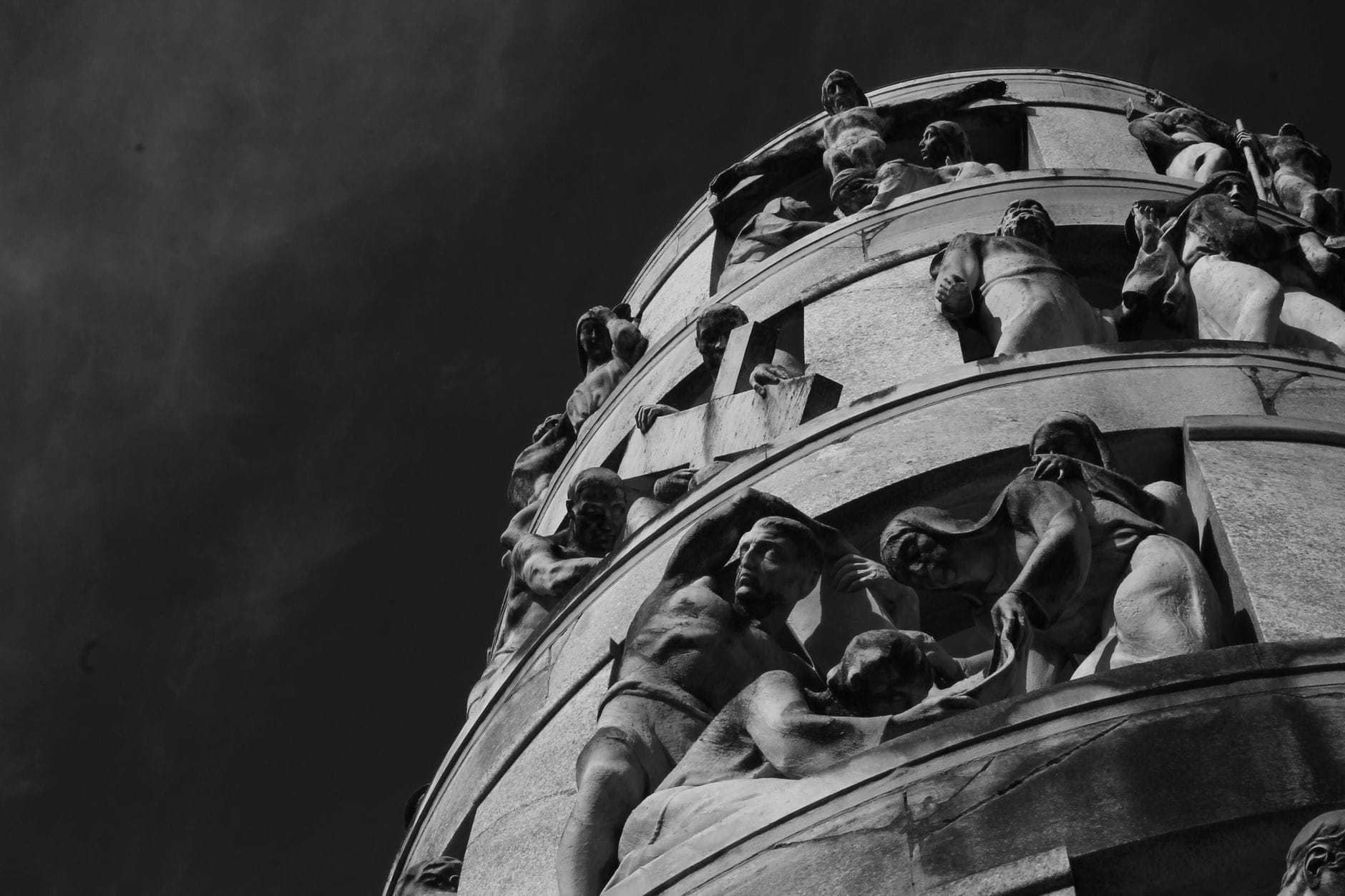 Suggestiva vista in bianco e nero dal basso verso l'alto di un grande monumento funebre circolare, ornato da sculture umane in movimento dinamico che si stagliano contro il cielo al Cimitero Monumentale di Milano.